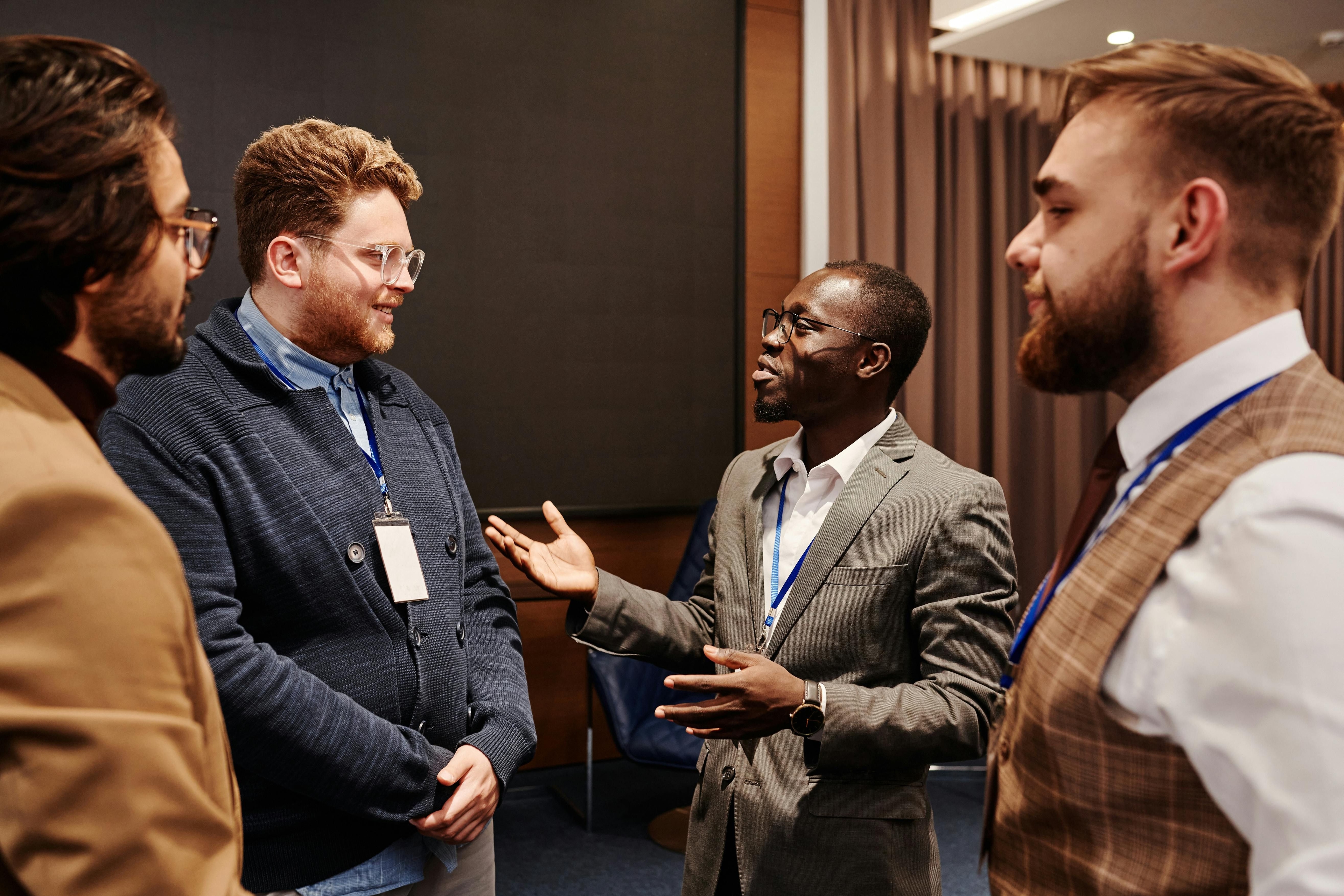 Grupo de quatro profissionais conversando em um ambiente de evento corporativo, com crachás de identificação. O cenário sugere uma oportunidade de networking e troca de ideias.