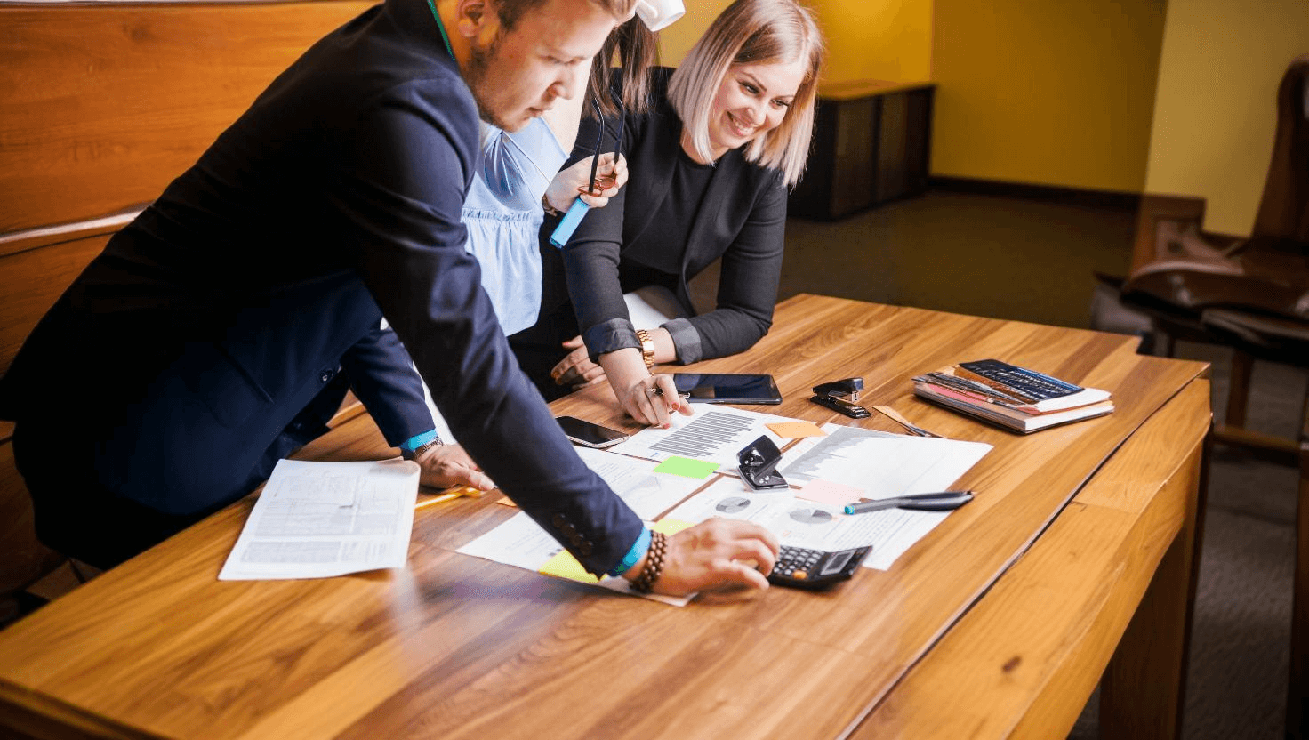 Grupo de profissionais analisando documentos e usando calculadoras em uma mesa de madeira durante uma reunião de trabalho. Foco em colaboração e produtividade. Felizes com a redução de custos da empresa.