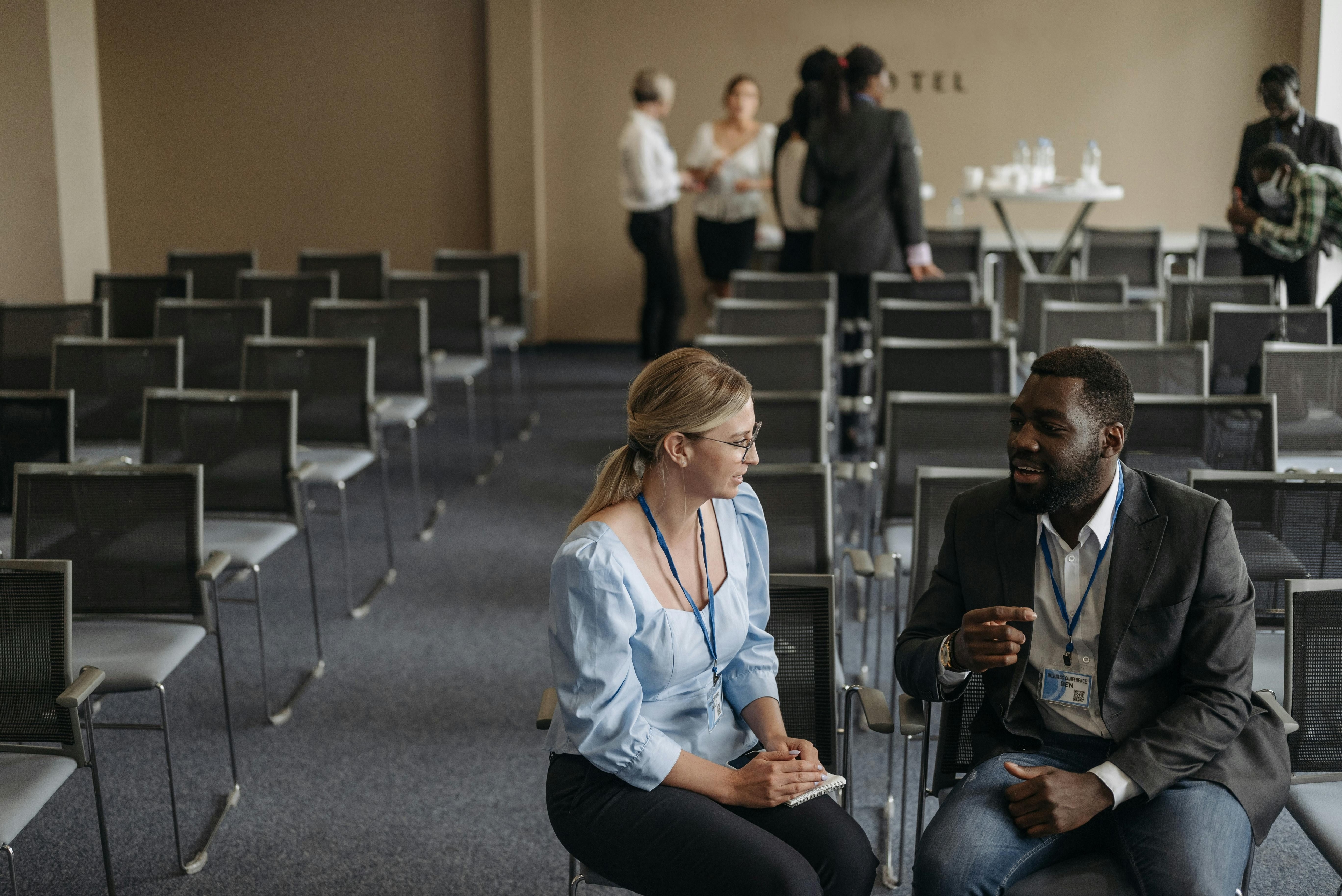 homem e mulher fazendo networking em uma sala de evento corporativo, pessoas ao fundo conversando