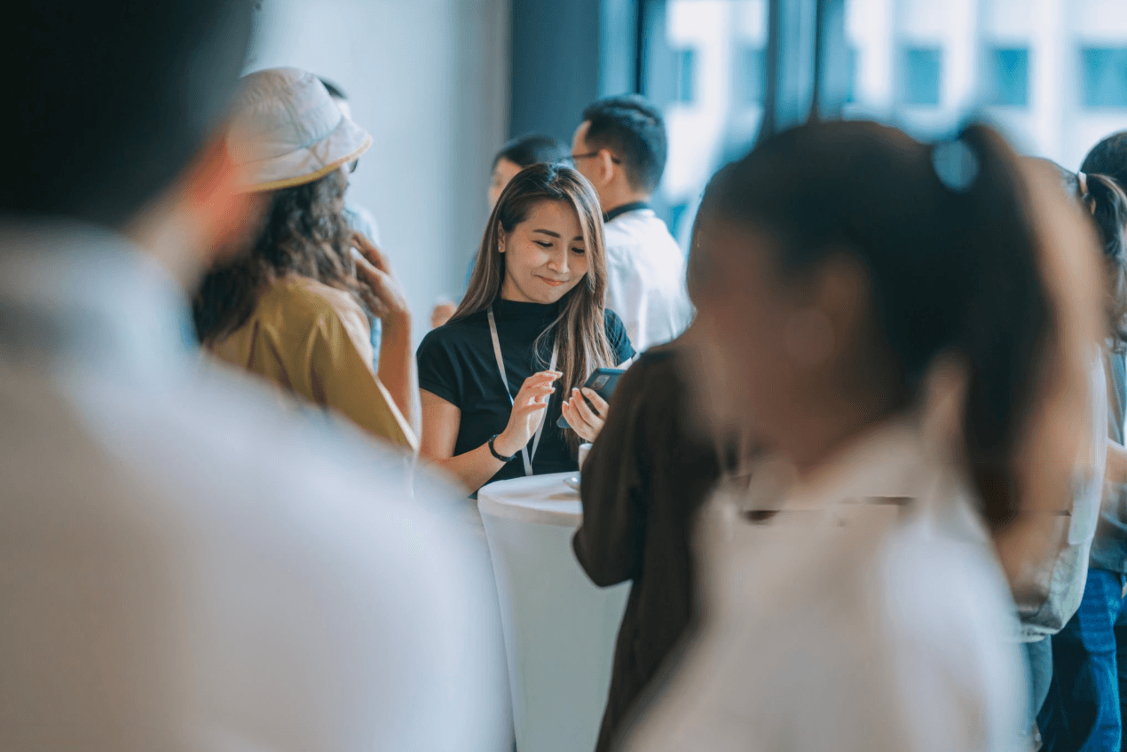 Pessoas em um evento de networking em um ambiente interno. No centro, uma mulher com cabelo liso e longo, introvertida, vestindo uma blusa preta, olha para o celular enquanto sorri. Ao redor, há outras pessoas desfocadas em conversas informais, algumas usando crachás de identificação. O ambiente tem uma atmosfera descontraída e profissional, com luz natural entrando por janelas grandes ao fundo.