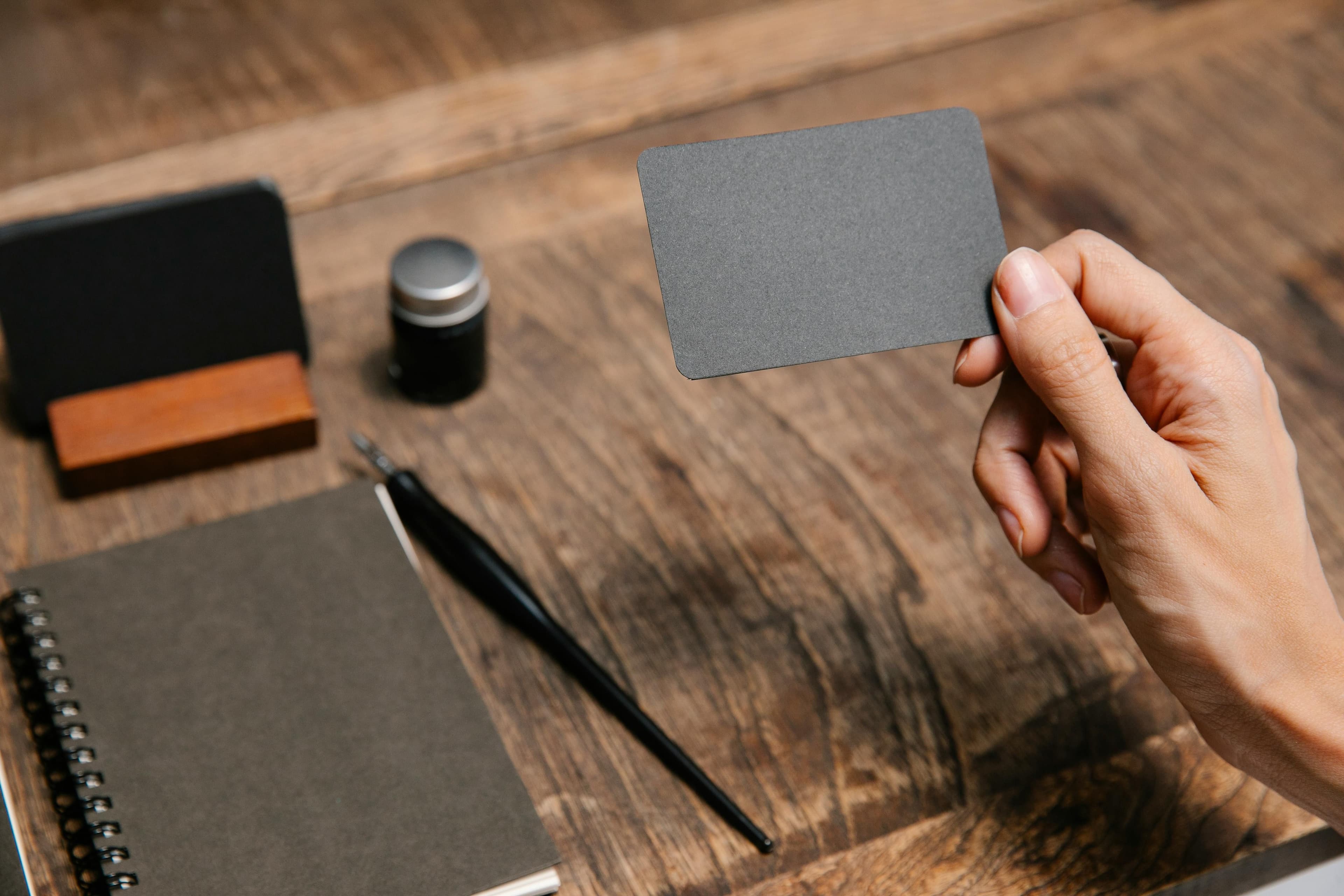 mano de mujer sosteniendo una tarjeta de visita negra frente a una mesa de madera con un cuaderno, bolígrafo y otros materiales